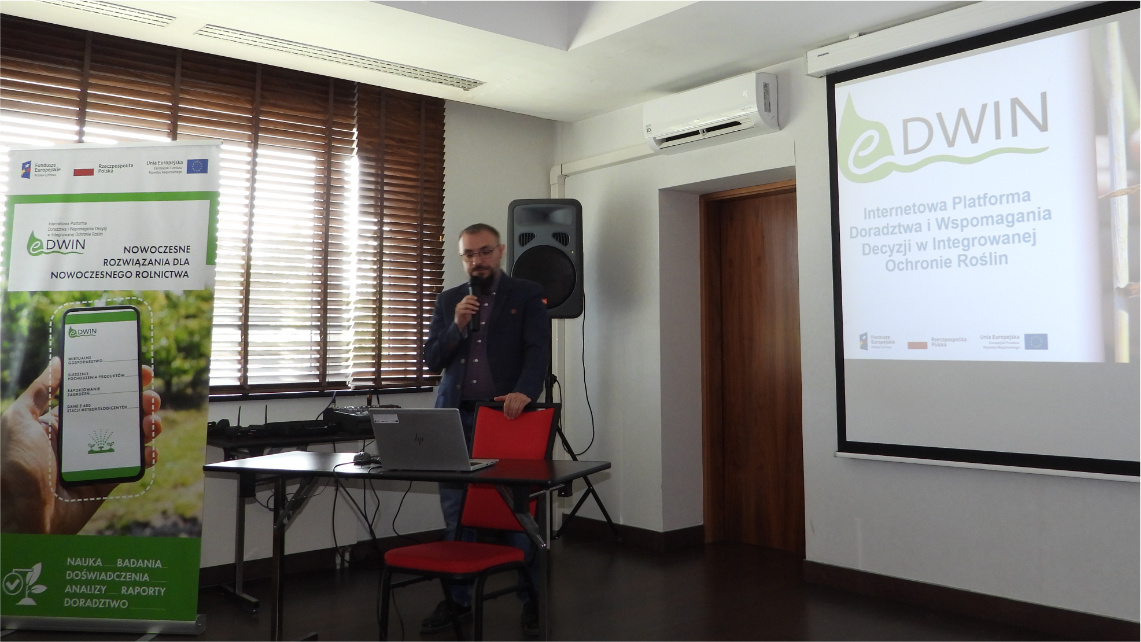 Maciej Zacharczuk, kierownik projektu eDWIN, podsumował trzyletnie prace nad platformą doradczą Wnętrze sali konferencyjnej. Młody mężczyzna - Maciej Zacharczuk - stoi z mikrofonem i wygłasza prezentację. Na telebimie widać prezentację. Obok mężczyzny stoi rollup eDWIN.