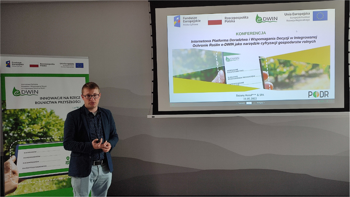 Daniel Dąbrowski z Pomorskiego Ośrodka Doradztwa Rolniczego w Poznaniu opowiadał o praktycznym wykorzystaniu Wirtualnego gospodarstwa w ramach aplikacji eDWIN Wnętrze sali konferencyjnej. Daniel Dąbrowski, młody mężczyzna w ciemnej marynarce, stoi przed rollupem promującym platformę eDWIN i wygłasza wykład. Po jego lewej stronie, na rzutniku widoczna jest prezentacja o eDWIN.