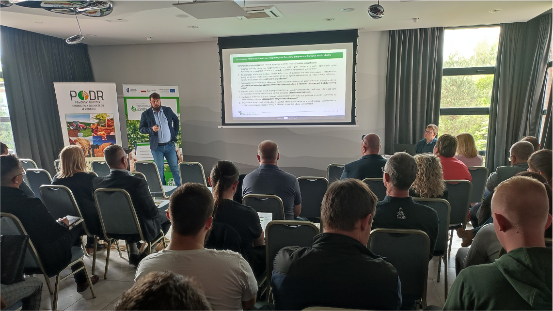 Marcin Baran z Instytutu Ochrony Roślin opowiadał o znaczeniu akcesu informacji w ochronie roślin Zdjęcie przedstawiające salę konferencyjną z widocznymi od tyłu słuchaczami, którzy siedzą na krzesłach i wysłuchują prezentacji Marcina Barana. Prowadzący widoczny jest z tyłu, obok niego jest rollup promujący eDWIN oraz rzutnik, na którym wyświetlona jest prezentacja.