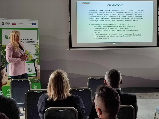 Anna Pietruczuk z Centrum Doradztwa Rolniczego w Brwinowie opowiadała o dalszych perspektywach rozwoju platformy doradczej eDWIN Młoda kobieta - Anna Pietruczuk - w różowej marynarce stoi i wygłasza prezentację. Za nią widać rollup promując platformę eDWIN. Kobieta patrzy się na siedzących przed nią słuchaczy. Z tyłu widoczna jest na rzutniku prezentacja.
