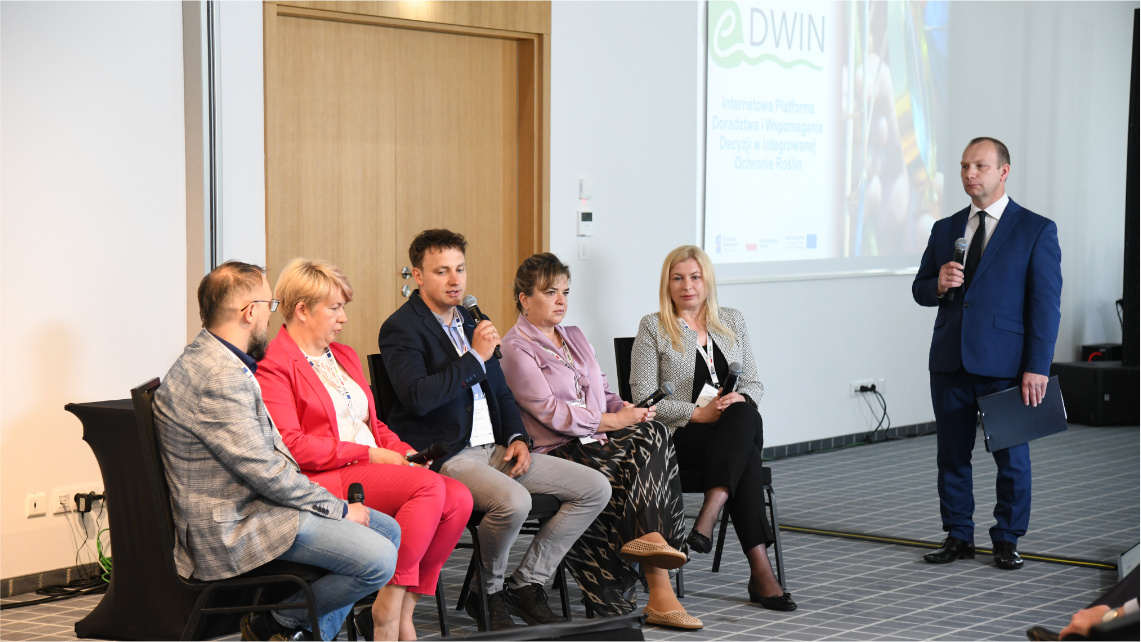 Końcowym elementem konferencji był panel dyskusyjny, w którym uczestniczyli wszyscy prelegenci. Grupa pięciu osób siedzi na krzesłach wewnątrz sali konferencyjnej. To uczestnicy panelu dyskusyjnego. Obok nich z mikrofonem w ręku stoi moderator w garniturze.