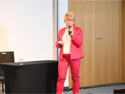 Magdalena Jakubowska z Instytutu Ochrony Roślin opowiadała o kwestiach dotyczących ochrony roślin Młoda kobieta w różowym garniturze - Magdalena Jakubowska - stoi z mikrofonem w ręku i wygłasza wykład. Za nią widoczny jest mały stół z czarnym obrusem.