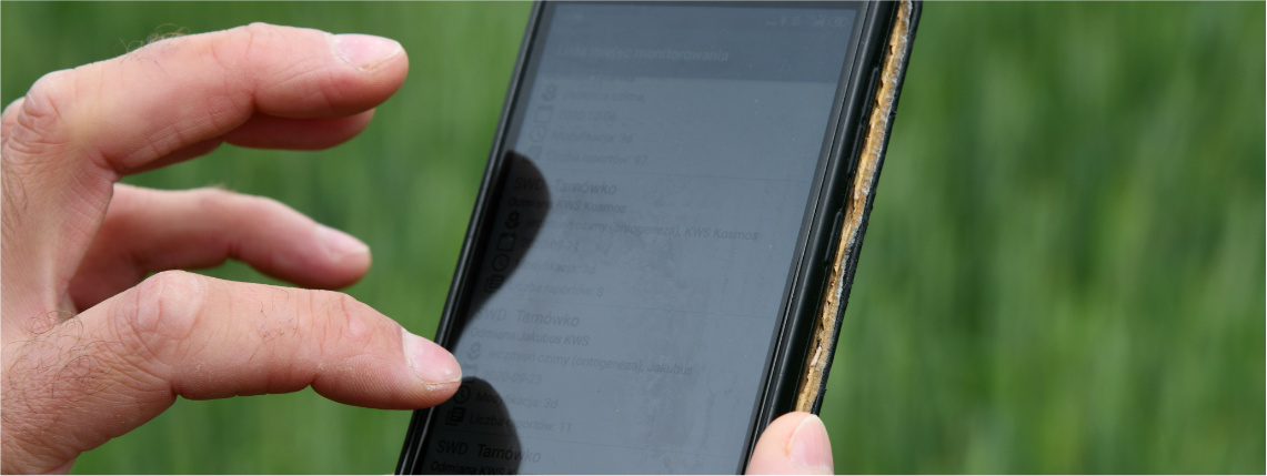 Prapremiera aplikacji eDWIN odbędzie się na czerwcowych targach w Sielinku. Oficjalna premiera będzie miała miejsce podczas Krajowych Dni Pola w Płońsku Na zdjęciu widać ekran telefonu, na którym wyświetlają się różne informacje. Dłoń dotyka ekranu telefonu. W tle widoczne zielone uprawy.