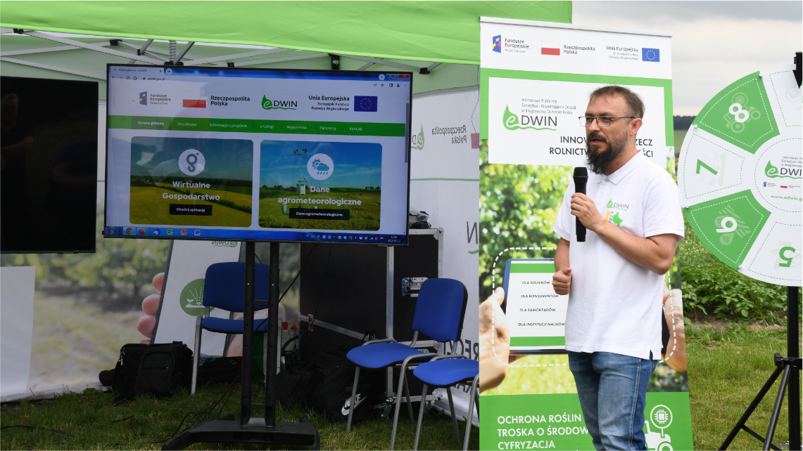 Lekcja polowa dla uczniów Kierownik projektu eDWIN Maciej Zacharczuk stoi koło stoiska platformy eDWIN oraz mówi przez mikrofon. W tle widać materiały promocyjne o eDWIN.