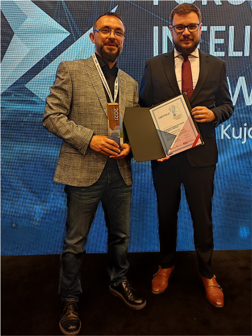 W imieniu WODR na Forum Inteligentnego Rozwoju byli informatycy Maciej Zacharczuk (po lewej) i Adam Fojud (po prawej) Maciej Zacharczuk (po lewej) i Adam Fojud (po prawej), informatycy WODR, stoją z nagrodą i dyplomem na scenie. Za nimi jest niebieskie tło.