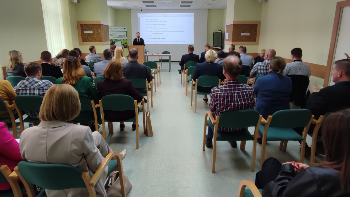 W konferencji uczestniczyło kilkadziesiąt zaproszonych osób Wnętrze pomieszczenia. Na zdjęciu widać od tyłu grupę kilkudziesięciu osób siedzących na krzesłach podczas konferencji oraz patrzących na prezentację na rzutniku, która jest przed nimi.