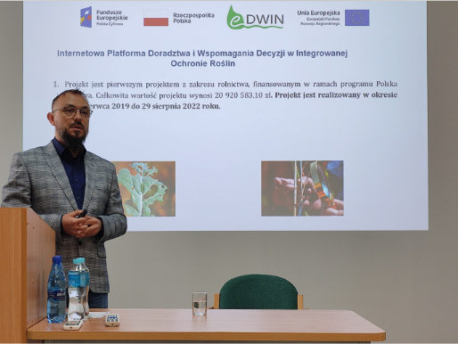 Kierownik projektu Maciej Zacharczuk, z Wielkopolskiego Ośrodka Doradztwa Rolniczego w Poznaniu, opowiadał o istocie projektu i platformy eDWIN Wnętrze pomieszczenia. Maciej Zacharczuk, mężczyzna w średnim wieku, stoi w marynarce przed rzutnikiem, na którym widoczna jest prezentacja o platformie eDWIN. Przed nim widoczny jest stół i zielone krzesło