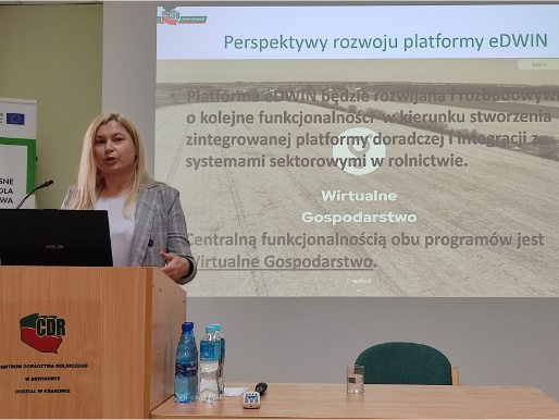 Anna Pietruczuk z Centrum Doradztwa Rolniczego opowiadała o perspektywach rozwoju platformy doradczej eDWIN Wnętrze pomieszczenia. Anna Pietruczuk, młoda kobieta, stoi w marynarce przed rzutnikiem, na którym widoczna jest prezentacja o Wirtualnym gospodarstwie. Przed nią jest mównica, na której stoi laptop.