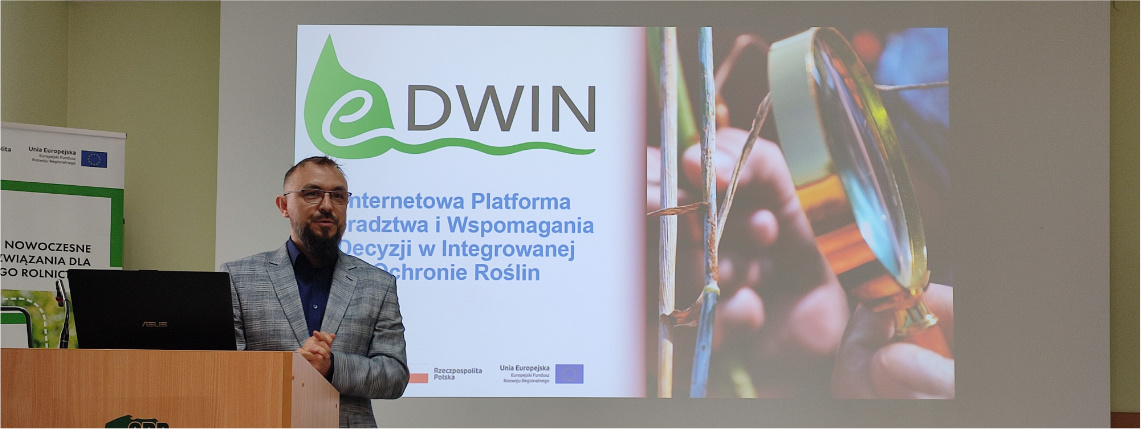 Kierownikiem projektu eDWIN jest Maciej Zacharczuk z Wielkopolskiego Ośrodka Doradztwa Rolniczego w Poznaniu Wnętrze pomieszczenia. Maciej Zacharczuk, mężczyzna w średnim wieku, stoi w marynarce przed rzutnikiem, na którym widoczna jest prezentacja o platformie eDWIN. Przed nim, na mównicy jest postawiony laptop.