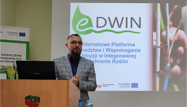Kierownikiem projektu eDWIN jest Maciej Zacharczuk z Wielkopolskiego Ośrodka Doradztwa Rolniczego w Poznaniu Wnętrze pomieszczenia. Maciej Zacharczuk, mężczyzna w średnim wieku, stoi w marynarce przed rzutnikiem, na którym widoczna jest prezentacja o platformie eDWIN. Przed nim, na mównicy jest postawiony laptop.