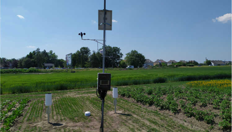 Usługa udostępnianie danych meteorologicznych byłaby niemożliwa bez kilkuset stacji meteorologicznych, które zostały zamontowane w całej Polsce. Słoneczny dzień. Na polu stoi stacja meteorologiczna, która zbiera dane meteorologiczne. W tle widać uprawy i drzewa.
